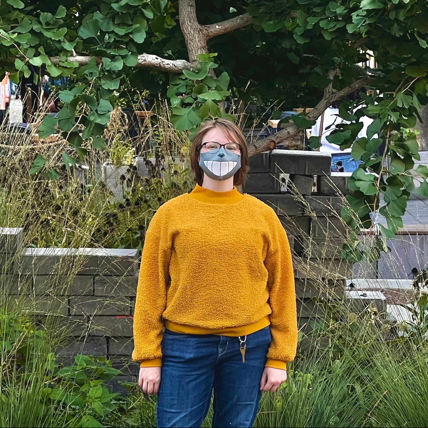 Misha standing under a gingko tree wearing a yellow sweater and Totoro Smile mask
